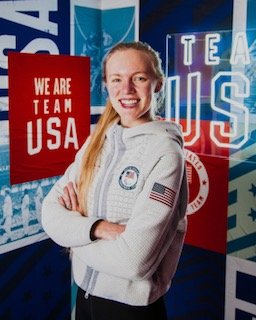 Jessica Heims has her arms crossed smiling with We are Team USA signs in the background